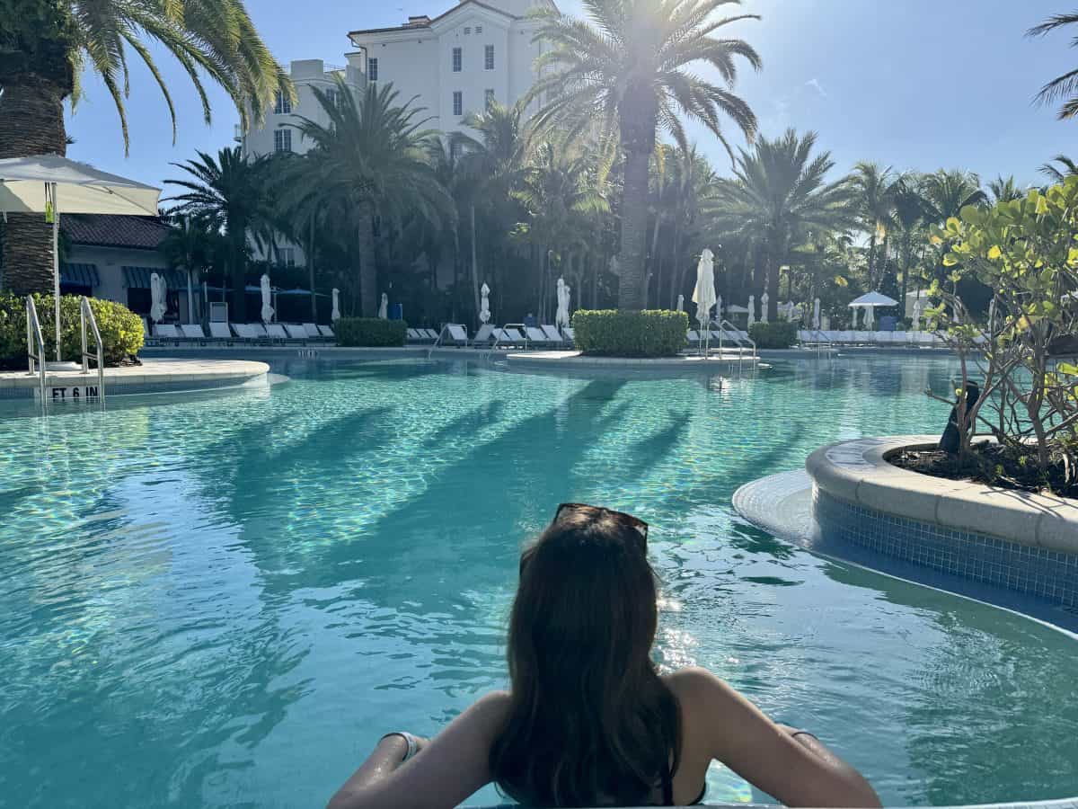 A woman with long hair relaxes in a clear, turquoise swimming pool surrounded by lush palm trees and sun loungers. The sun shines brightly, casting reflections on the waters surface. A white building is visible in the background.