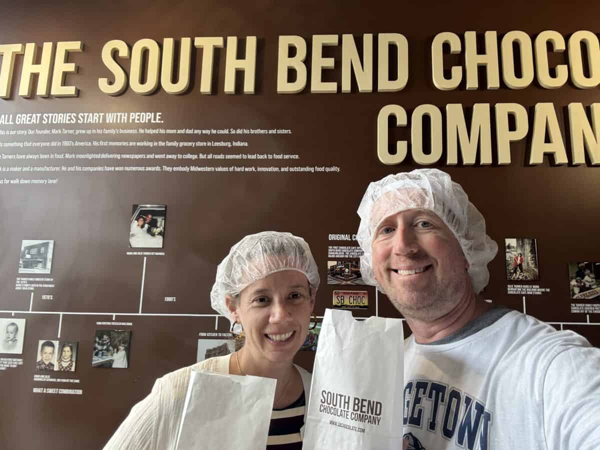 two people wearing hairnets posing with a bag of chocolate in front of the timeline of the South Bend Chocolate Factory in South Bend, IN